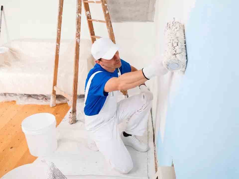 Peintre en chemise bleue et salopette blanche appliquant un rouleau sur un mur bleu clair à l'intérieur