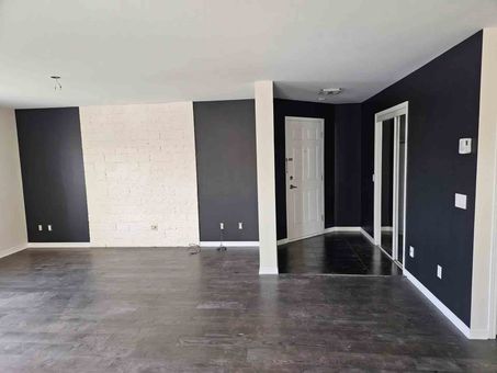 Salon moderne vide avec des murs d'accent sombres, une cheminée en briques blanches et un parquet gris.