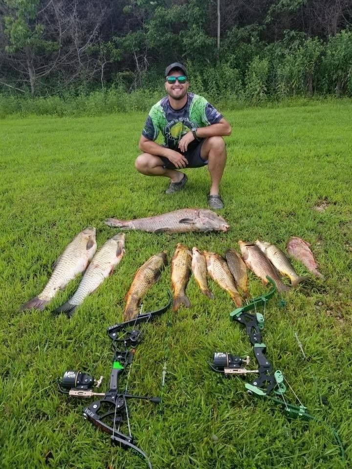 Man with fish and bows on grass. He's smiling. Green and brown tones, outdoors.