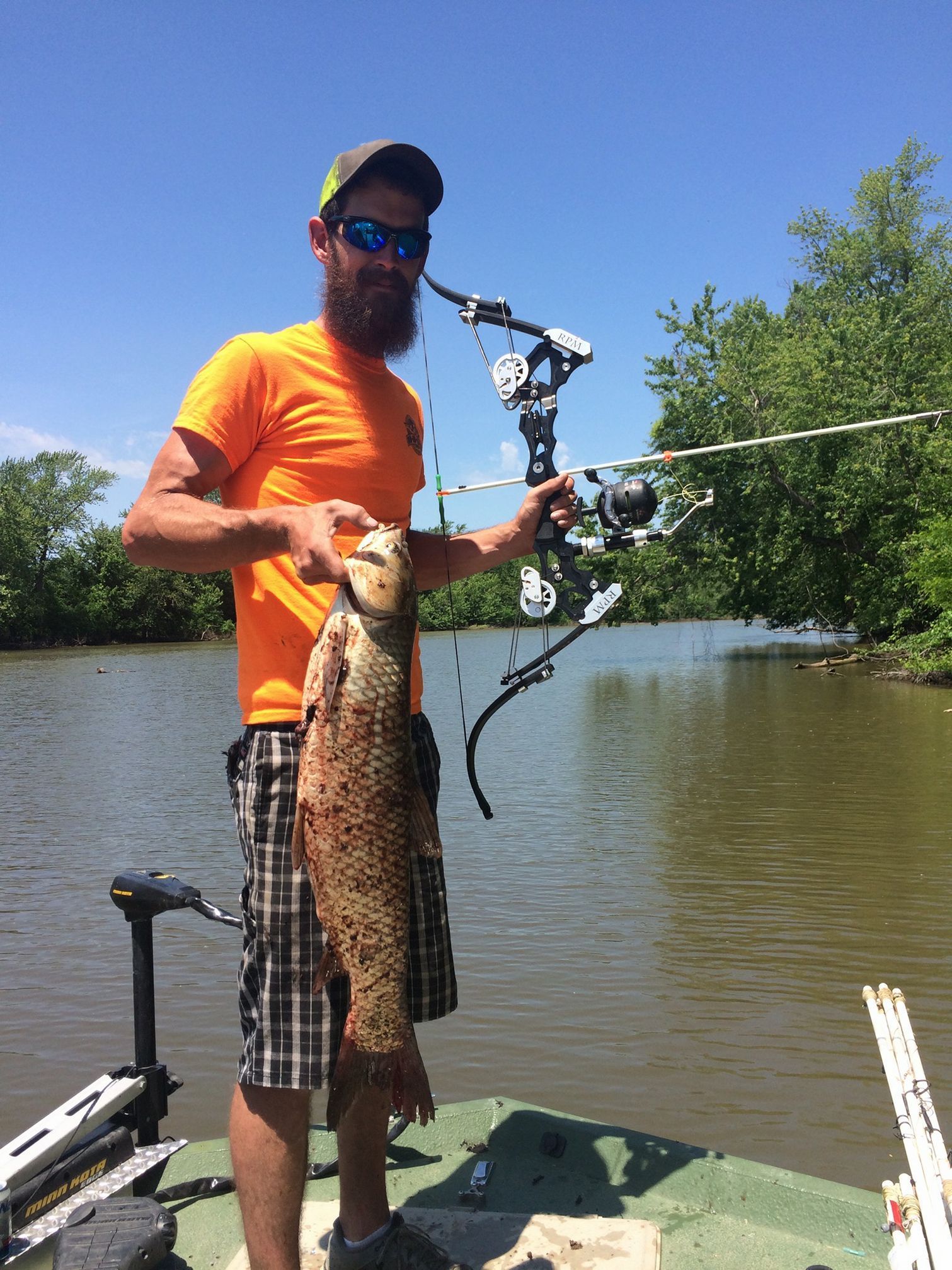 Man in orange shirt on boat holding large fish and bow. River setting, sunny.