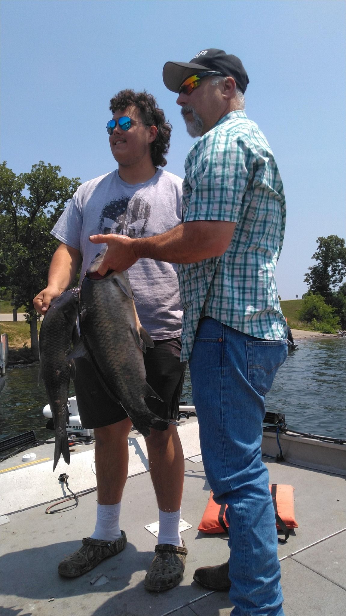 Two people on a boat display large fish they caught. One points toward the fish. Sunny day.