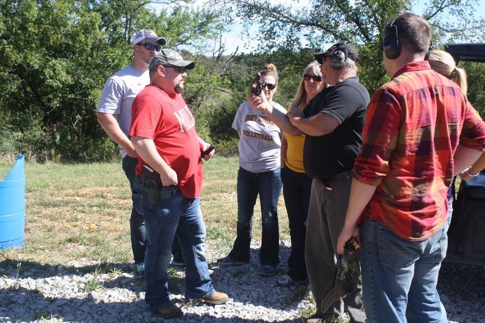 A group of people at an outdoor shooting range. A man in a red shirt gestures while others observe.