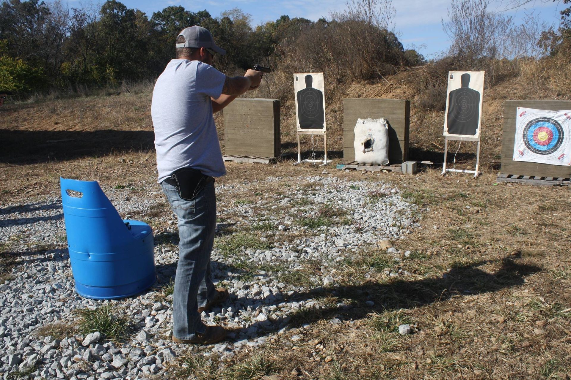 Man aiming a handgun at paper targets at an outdoor shooting range on a sunny day.