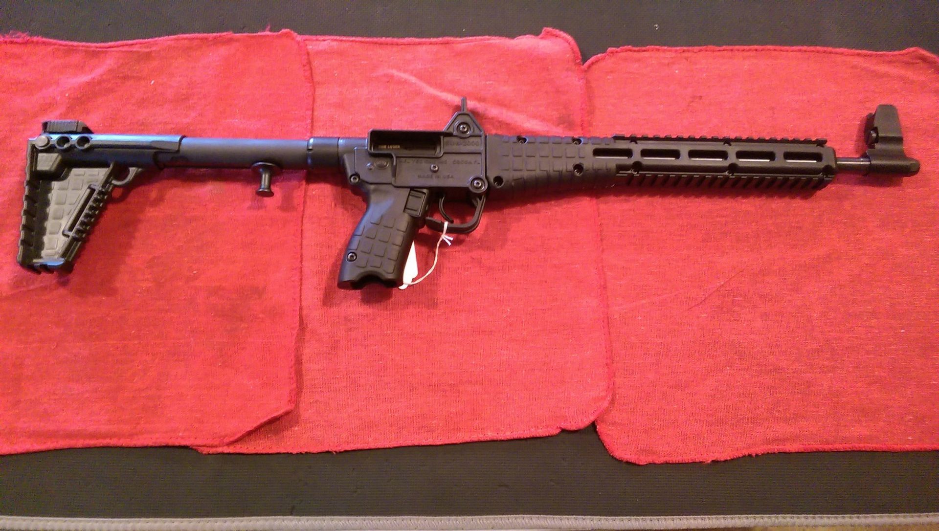 Black semi-automatic rifle with folding stock and tactical rail, resting on a red surface.