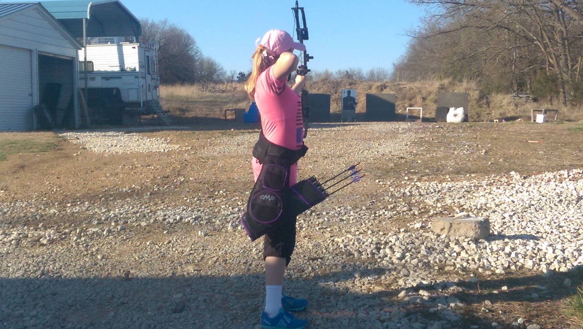 Young person in pink shirt aiming bow and arrow at target outdoors.