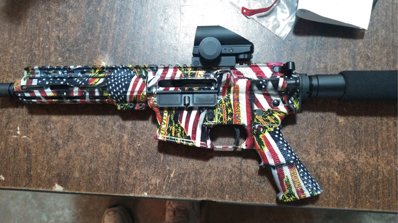 AR-15-style rifle, American flag camouflage pattern, red dot sight, black suppressor, lying on a wooden surface.