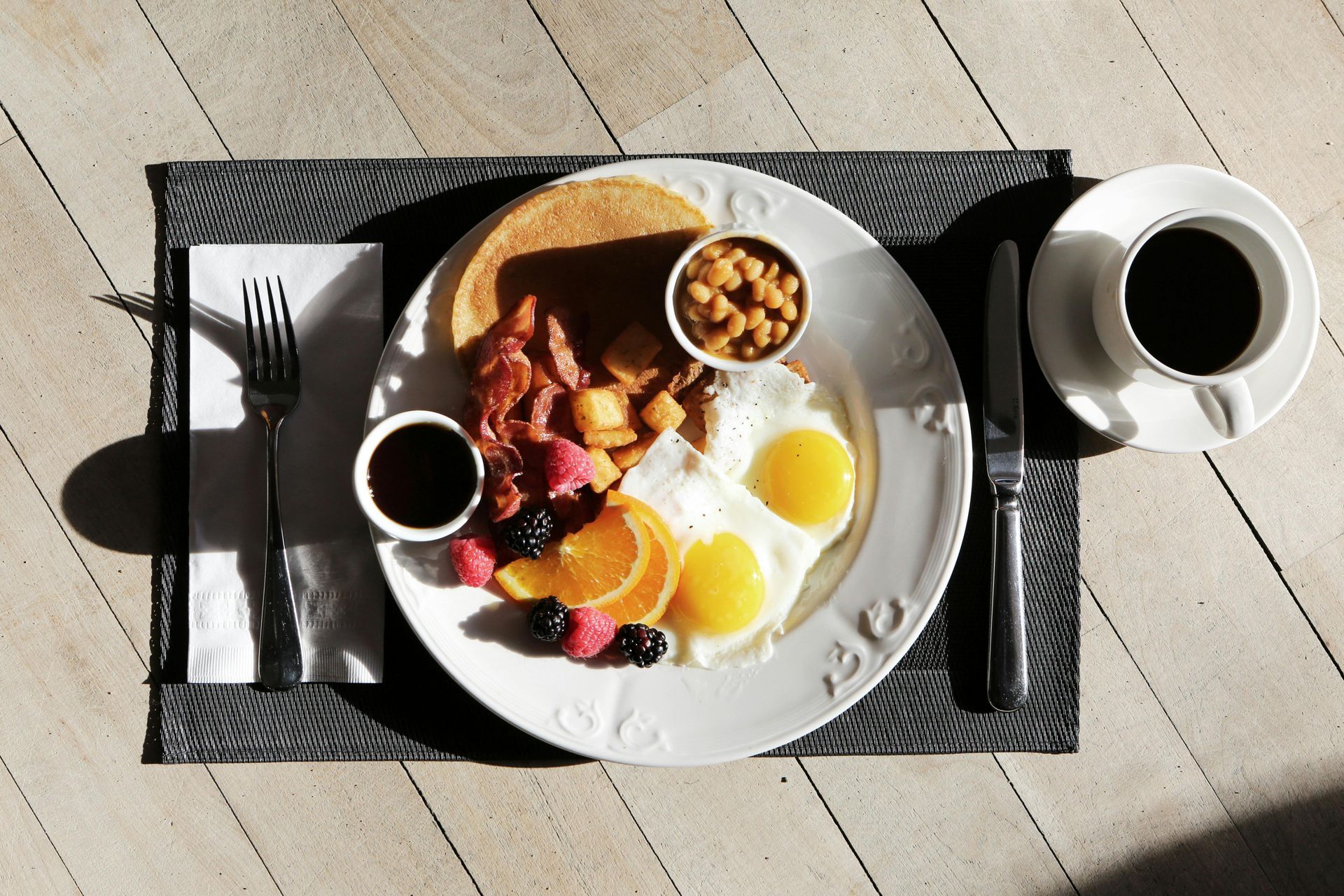 Breakfast plate with eggs, pancakes, bacon, fruit, and a cup of coffee.