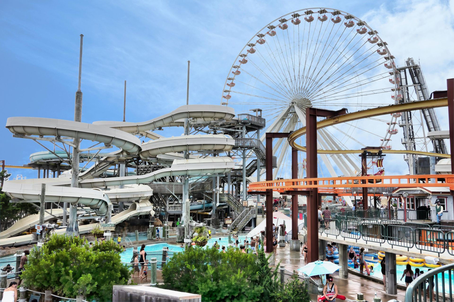 Water park with slides and a Ferris wheel on a sunny day. People in pools and on walkways.