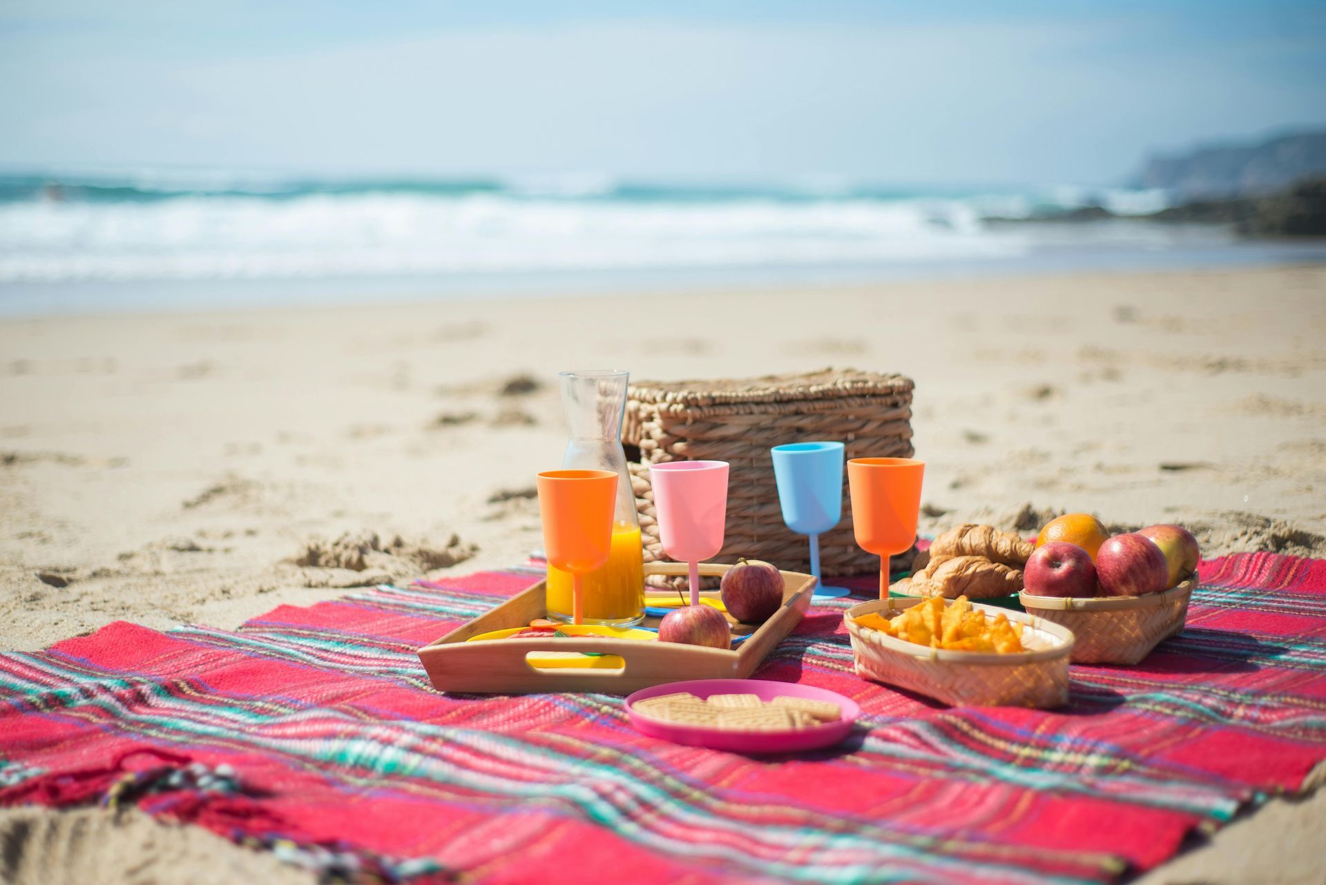 Picnic setup on a sandy beach: basket, glasses, fruit, chips on a red plaid blanket near the ocean.