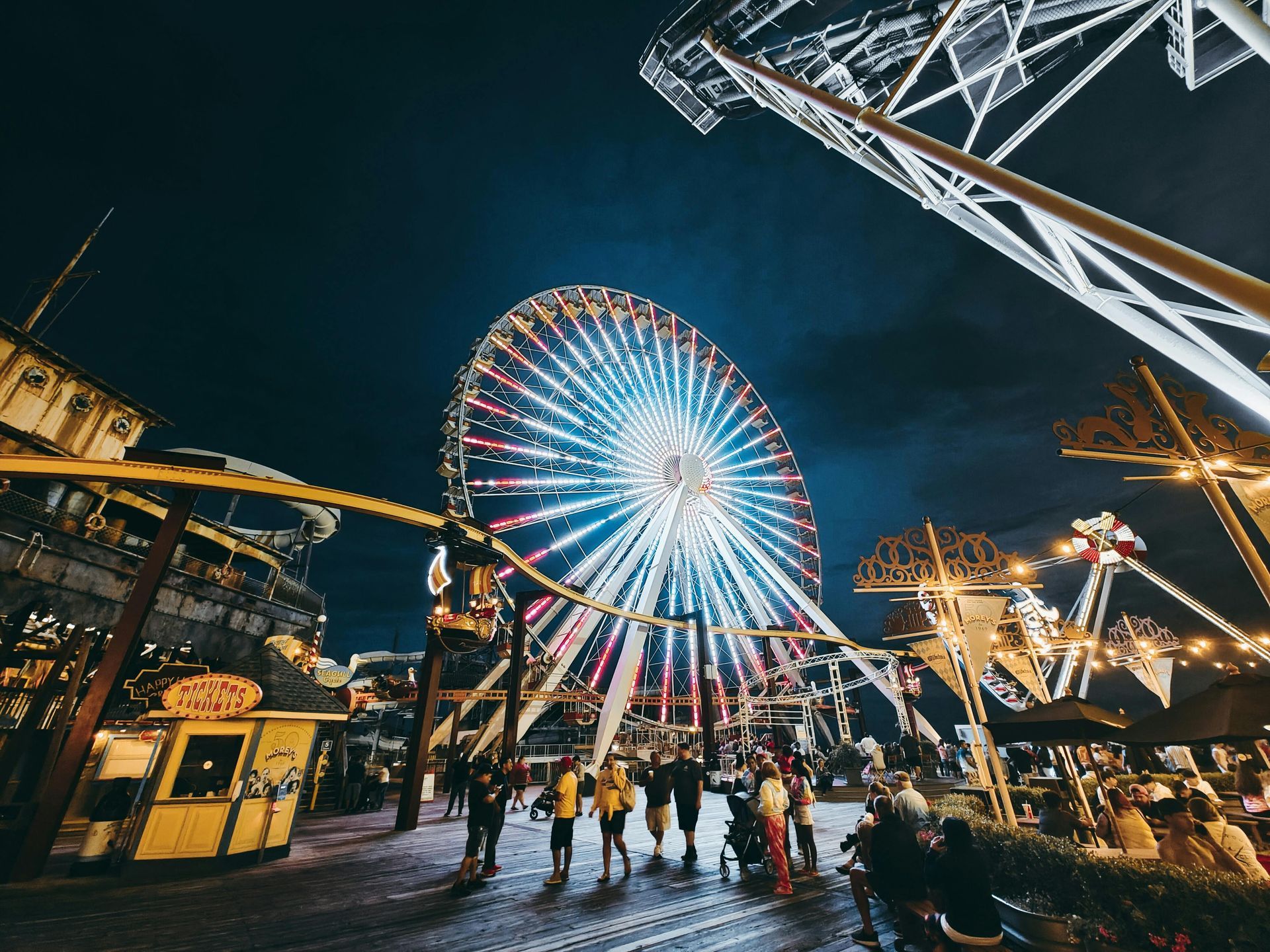 Nighttime amusement park with a brightly lit Ferris wheel; people walk on a boardwalk.