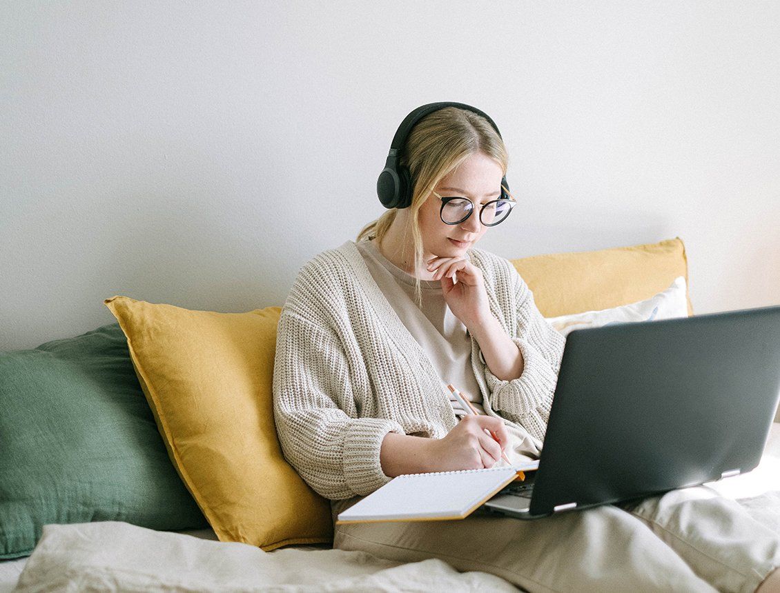 Image of female wearing headphones sitting on couch with laptop writing