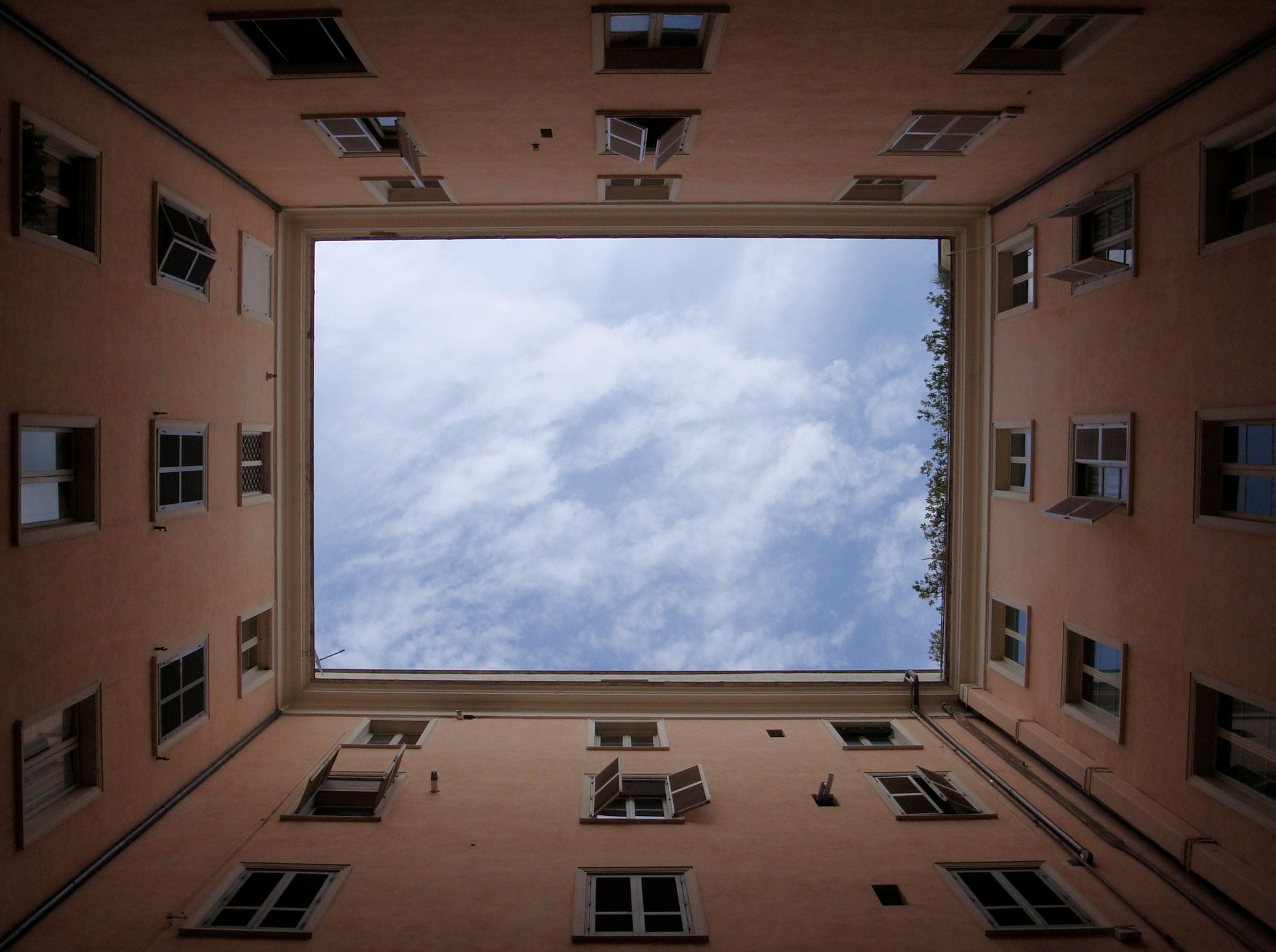 Looking up at the sky through a square courtyard. Pale pink walls with many windows. Blue sky and clouds.