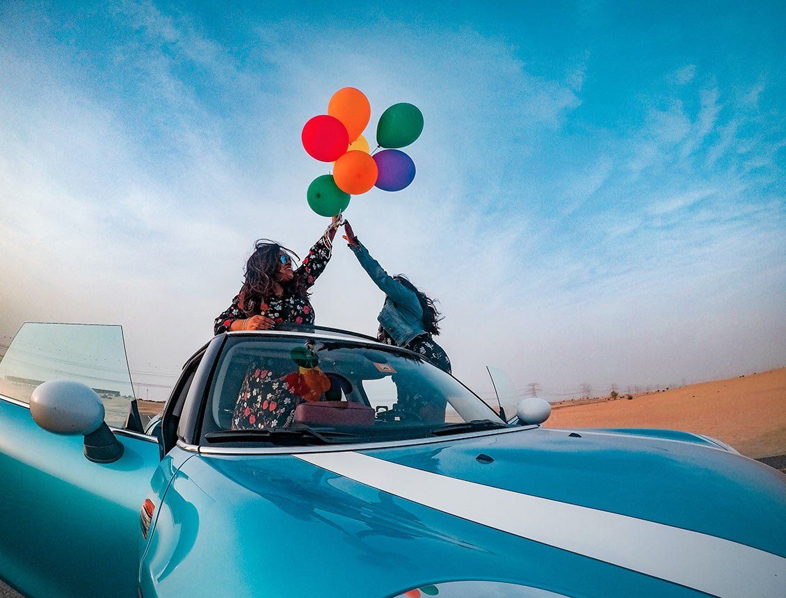 low angle wide shot of two people standing up on convertible car holding baloons