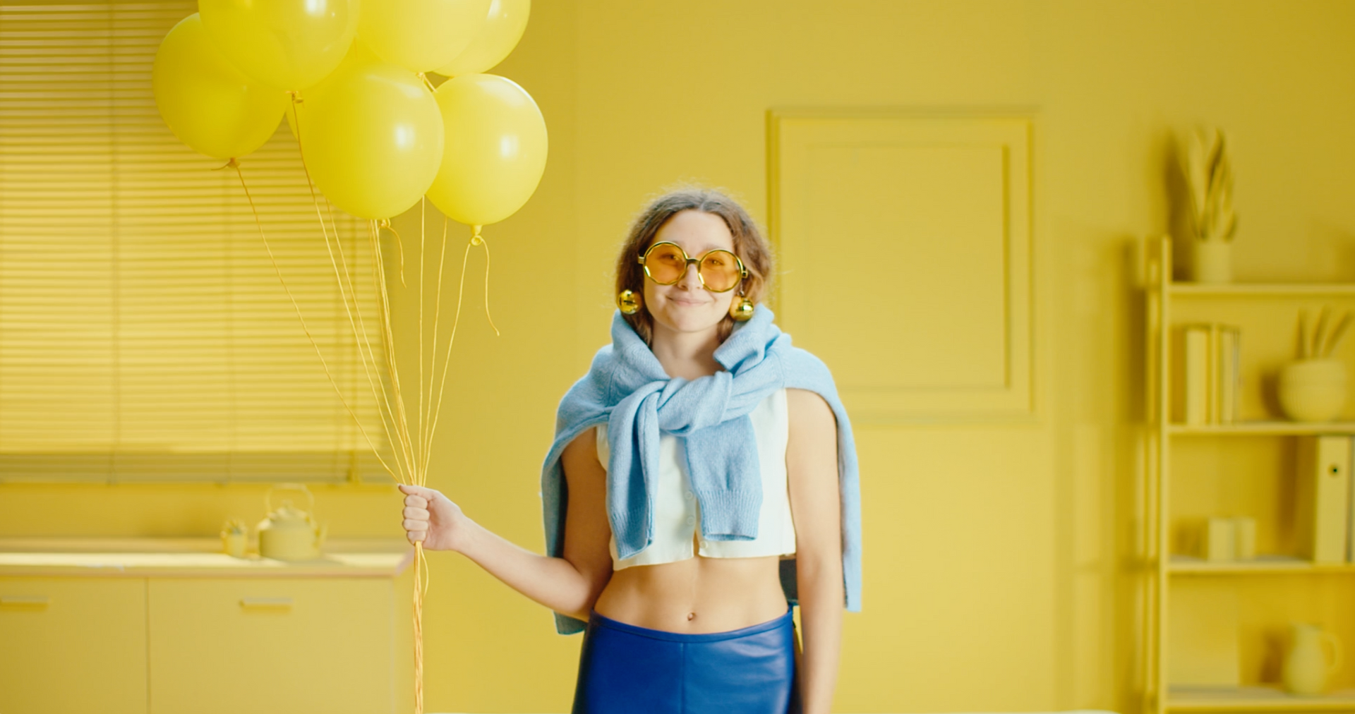 Woman in yellow room holding balloons; she wears blue, a sweater, and sunglasses.