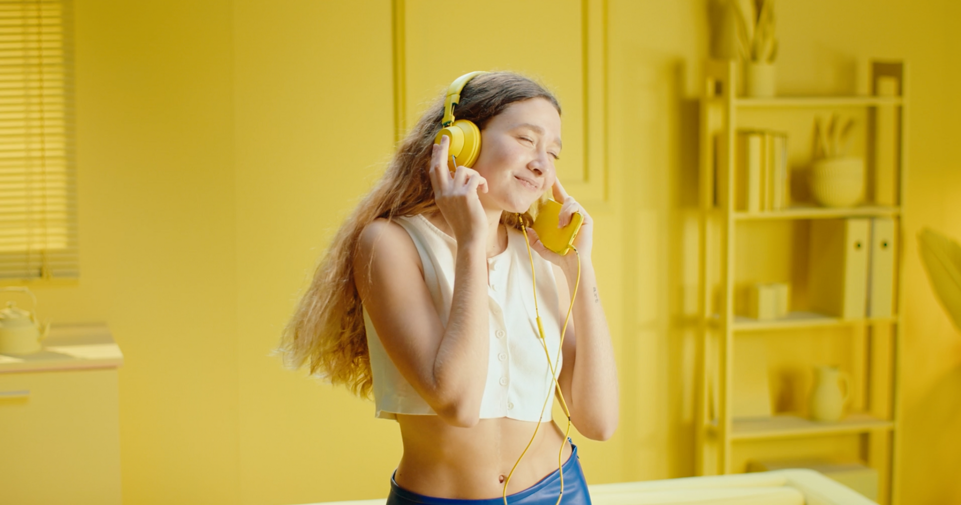 Woman in a yellow room with yellow headphones, smiling and listening to music. She wears a white crop top and jeans. Woman in a yellow room with yellow headphones, smiling and listening to music. She wears a white crop top and jeans.