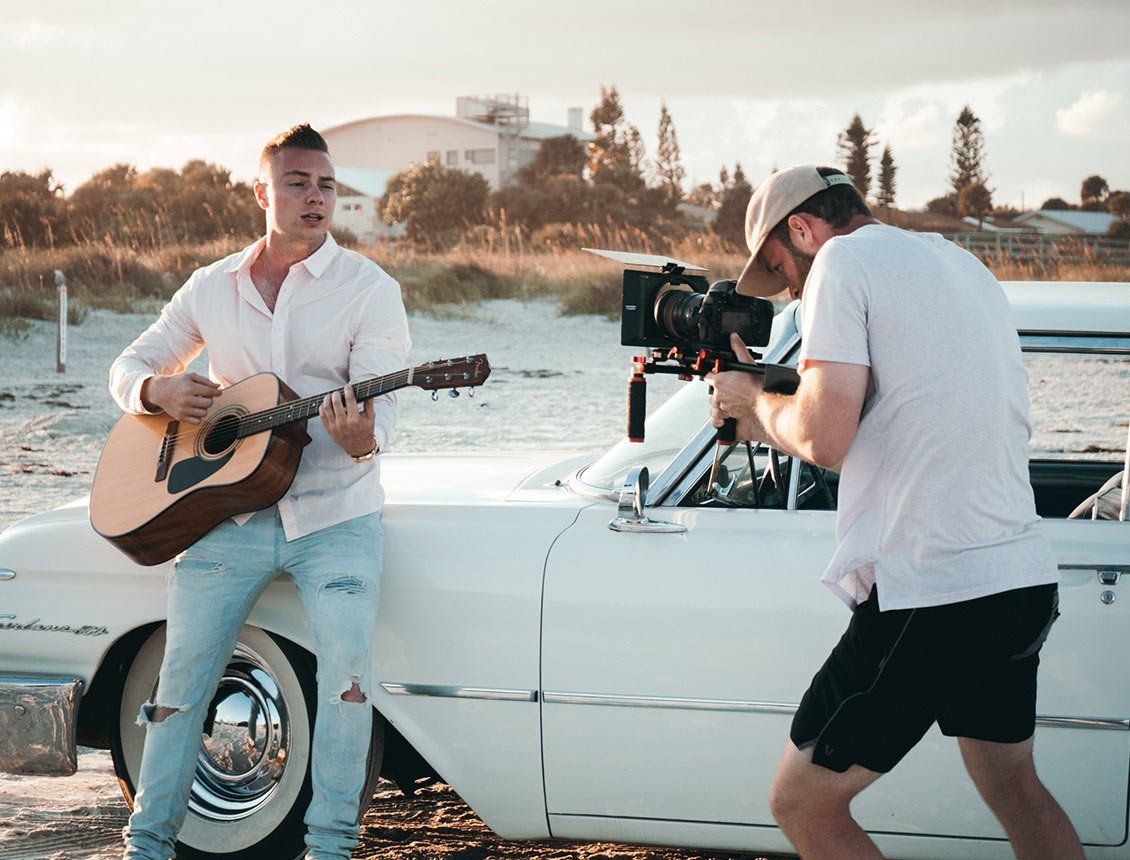 behind the scenes image of a videographer filming a man playing a guitar in front of car