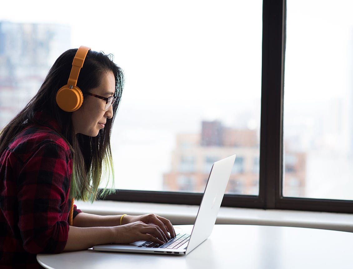 Image of woman wearing yellow headphones working on laptop in front of office building window