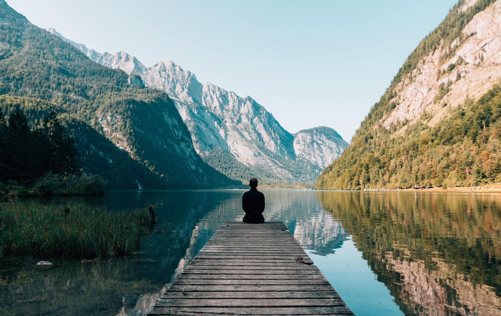 Image of a man sitting at end of dock with mountains and lake in front of him.