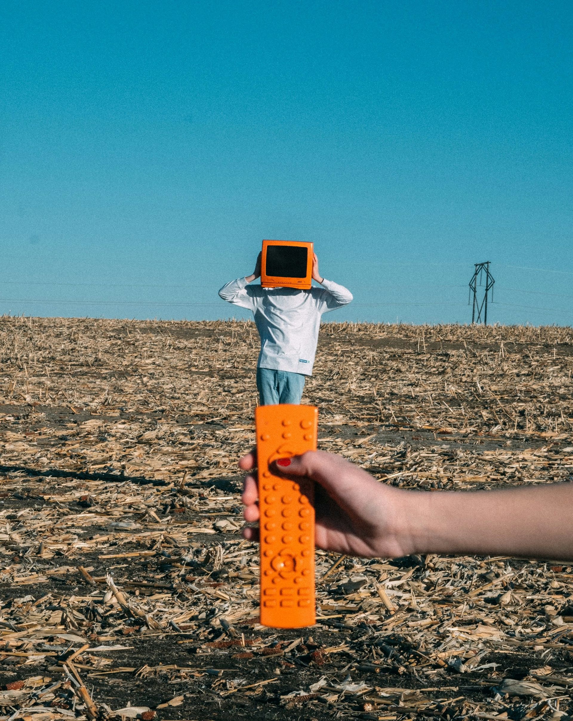 Person with a retro TV for a head, hands up. Orange remote held in front. Brown, textured background, blue sky.