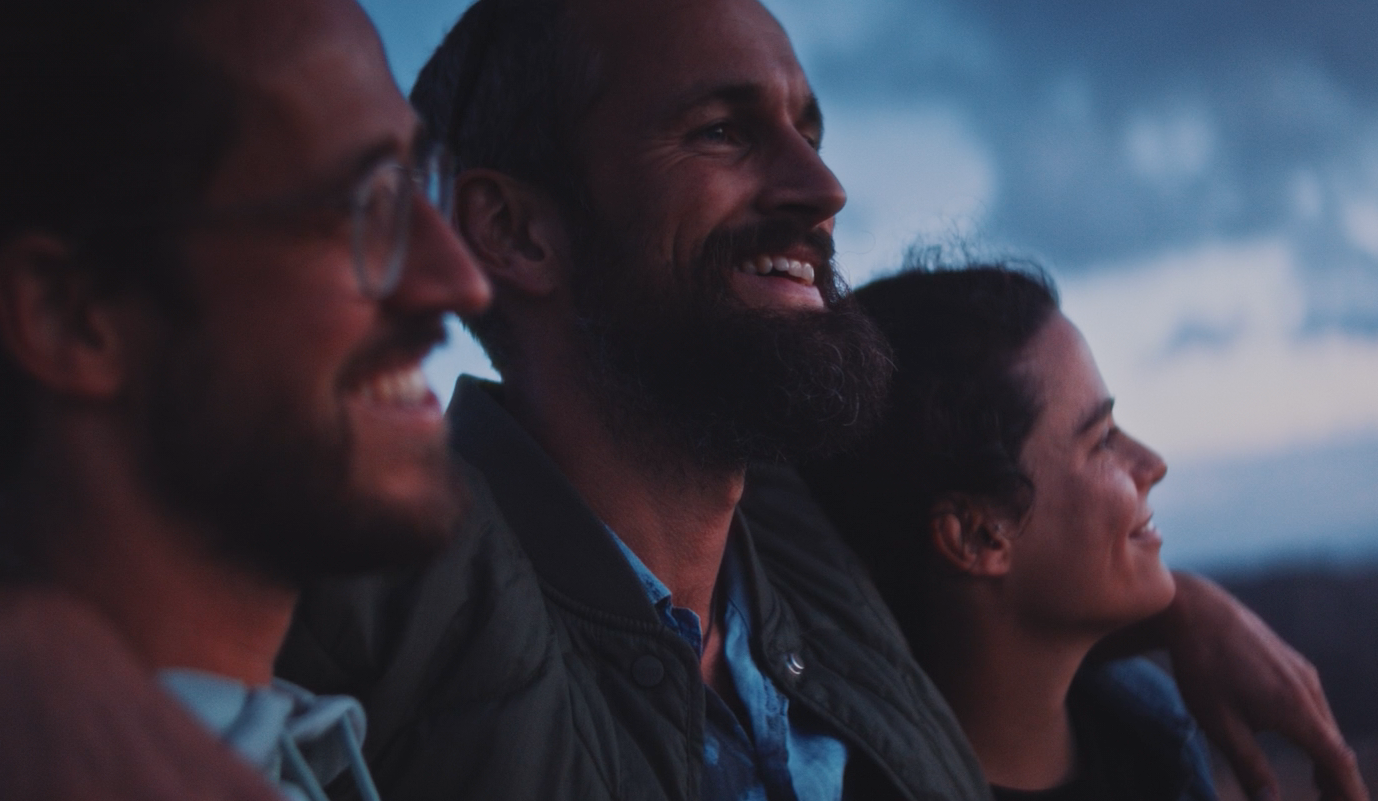 Three people smiling and looking upwards. Blue sky backdrop. Man has arm around woman.