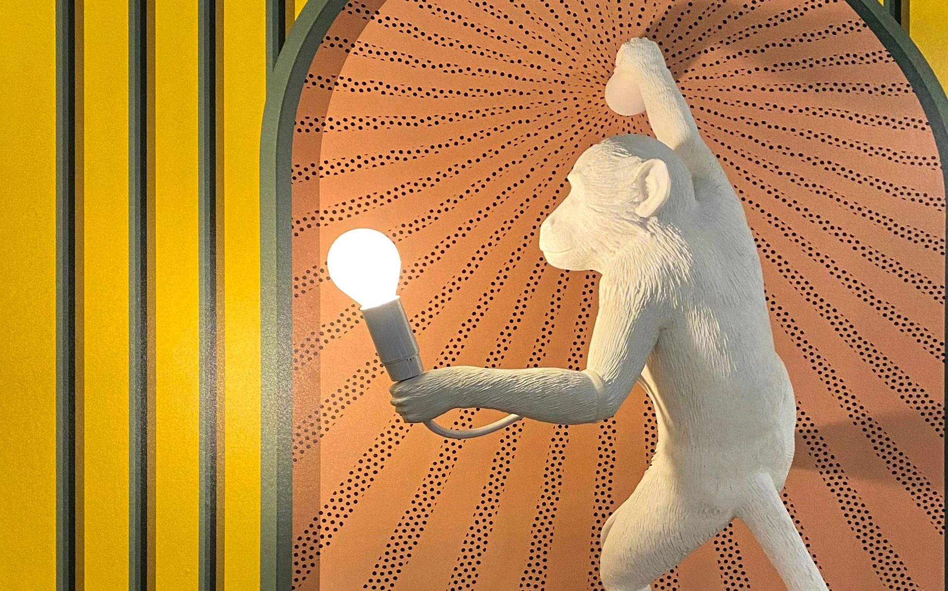 White monkey figure holding a lit bulb against a radiating pattern within an archway, on a striped background.