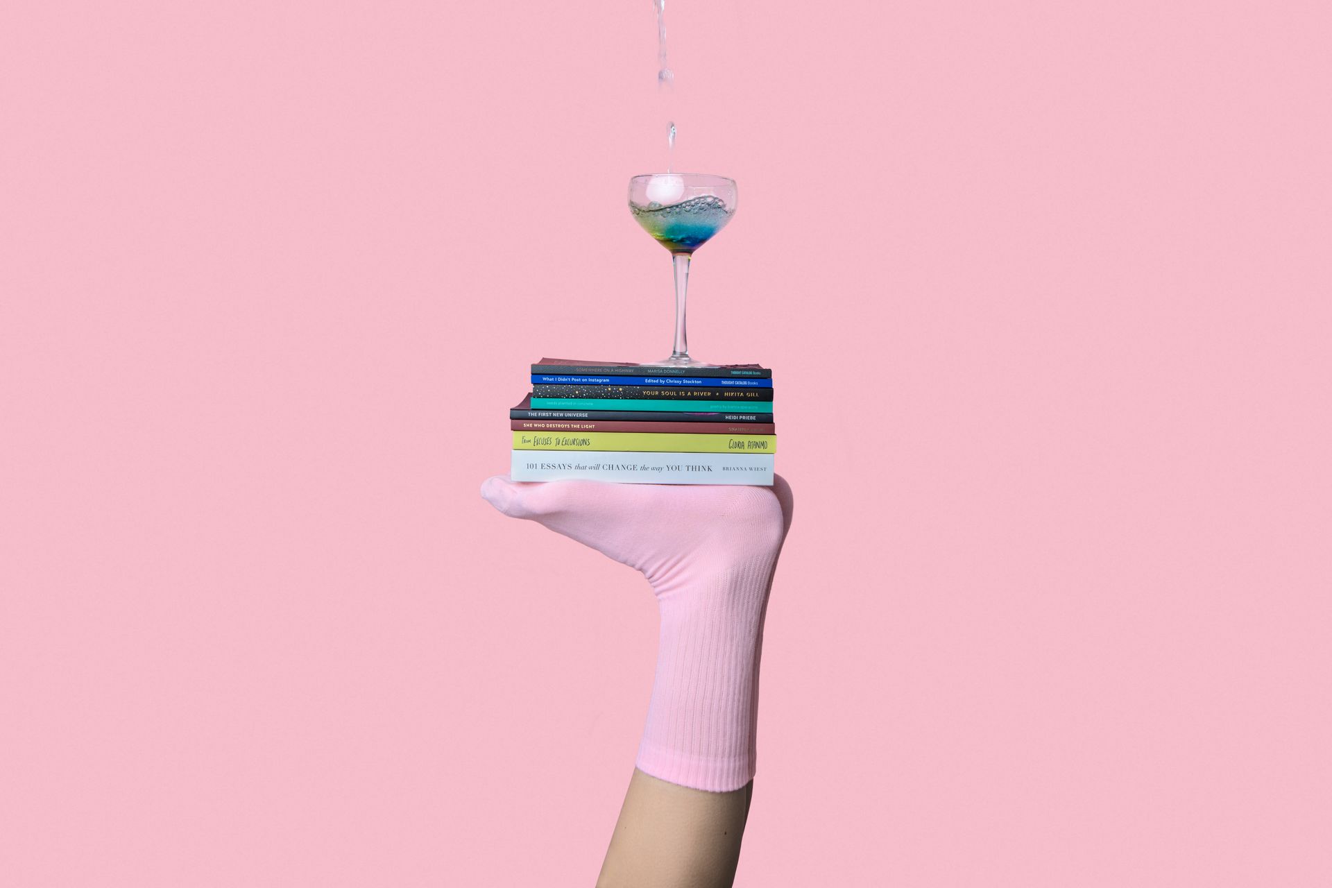 Feet balancing books with a cocktail glass on top over pink background