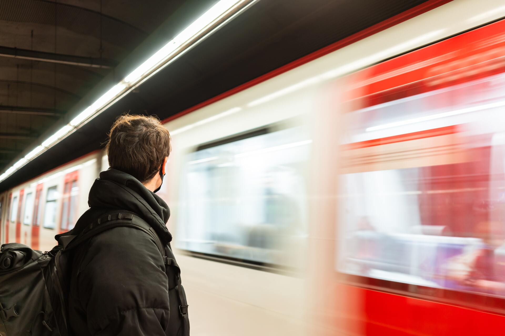 Image from behind of man wearing a mask at subway platform as a subway goes by in motion blur