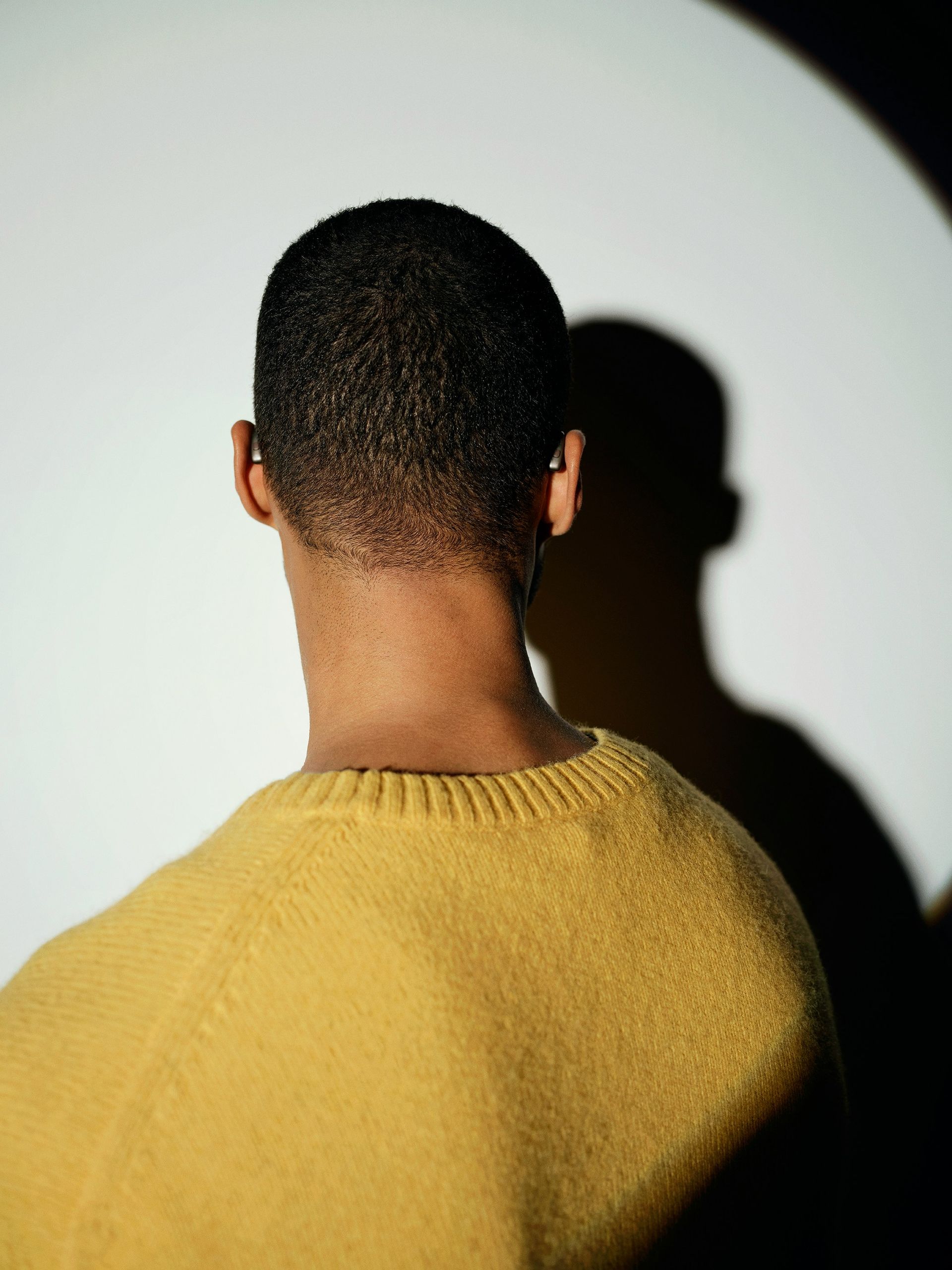 Person wearing a yellow sweater, viewed from behind, in front of a white wall with a circular shadow.