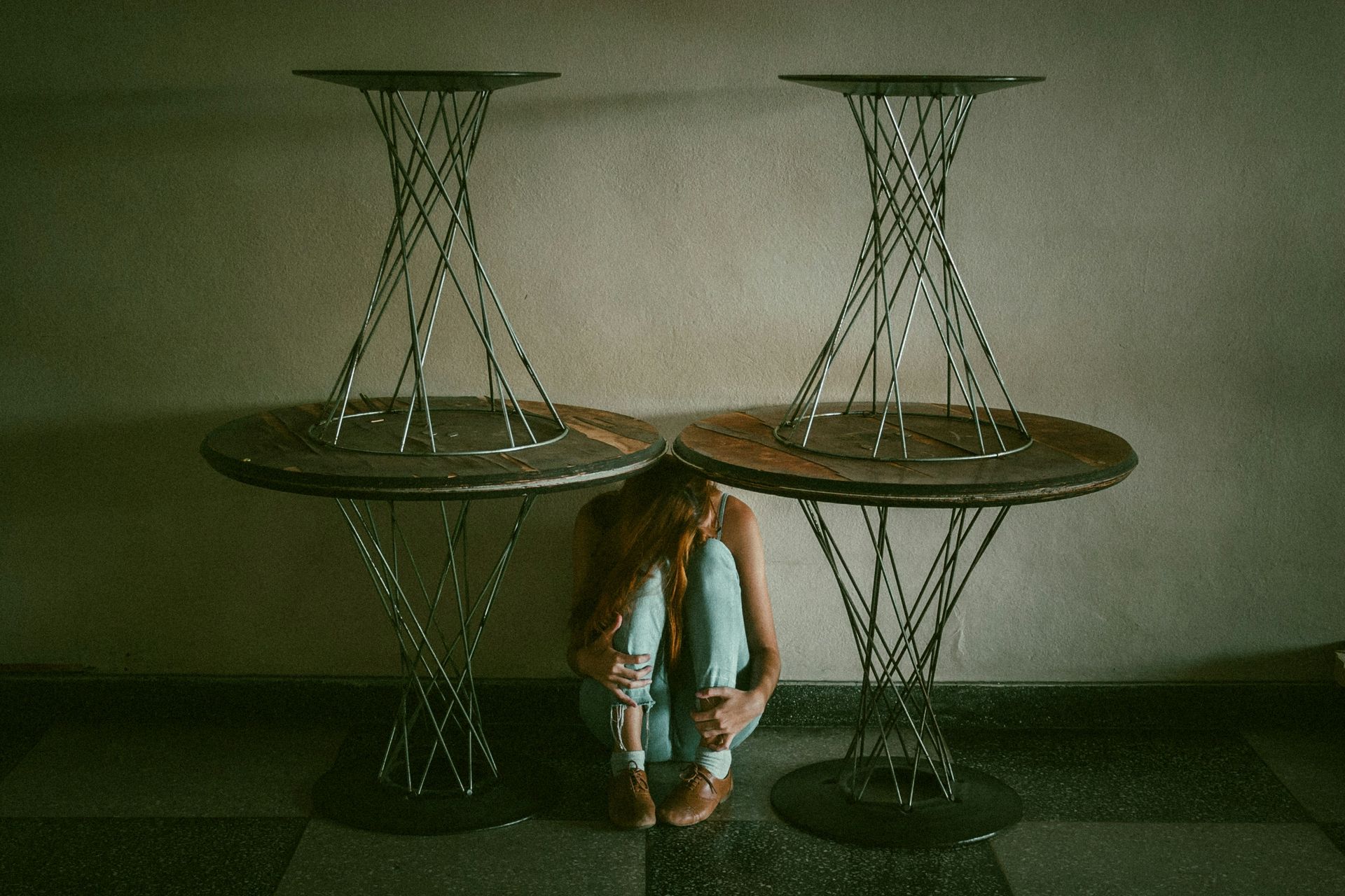 Person crouched between two spiral-legged tables; interior setting, neutral tones.