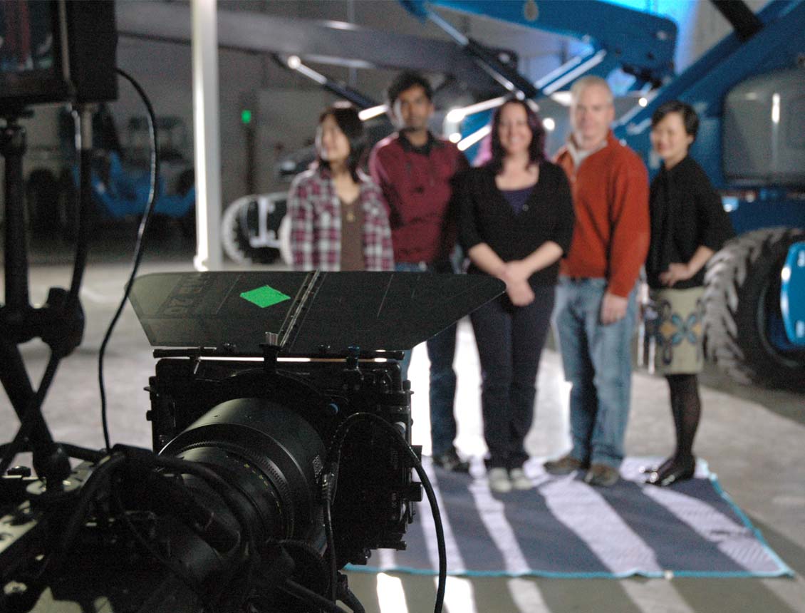 Behind the scenes wide shot of an Arri Alexa camera in the foreground and on-camera video subjects in the background, standing in front of large Genie lift equipment. Spin Creative video production shoot in Seattle, WA for a brand film for Genie.