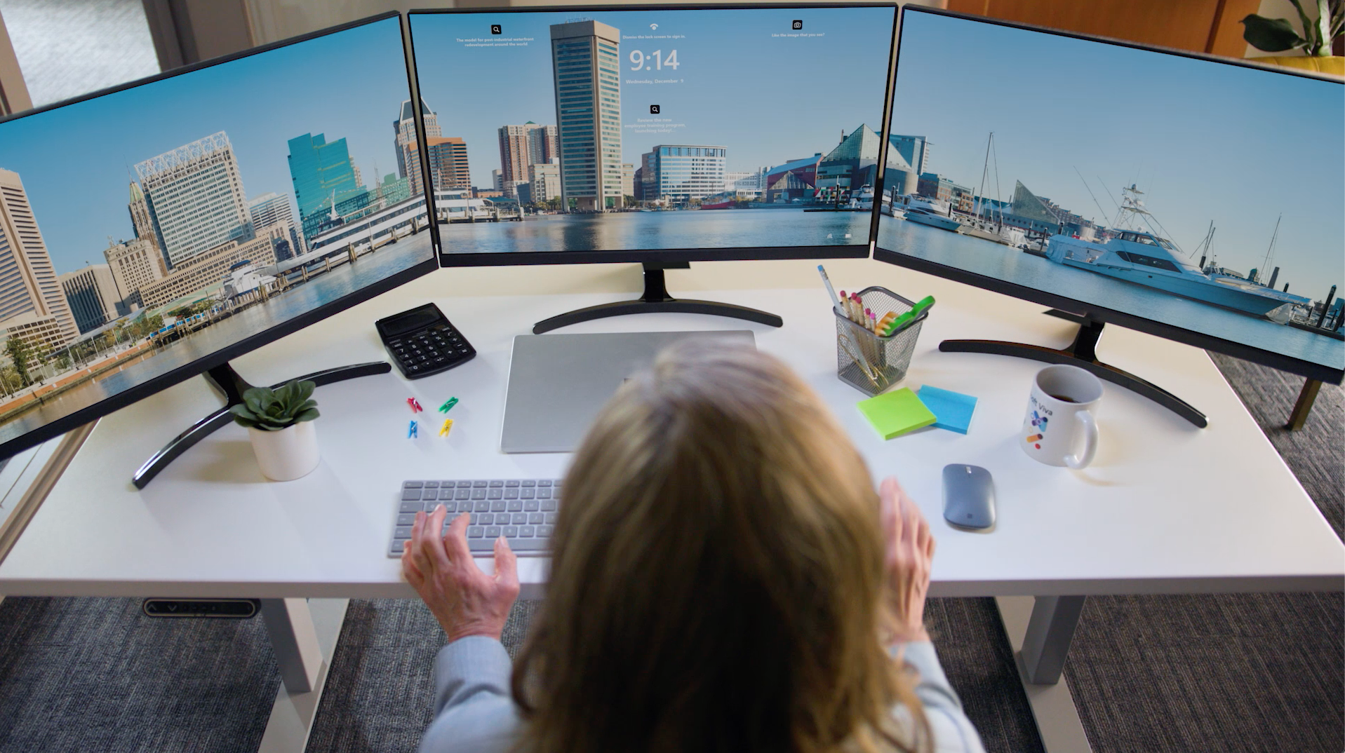 A woman is sitting at a desk with three computer monitors on it.