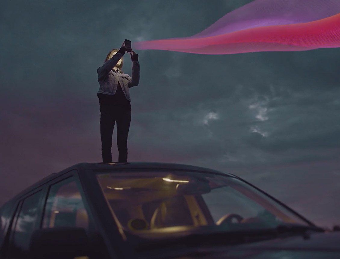Image of woman at night on top of car looking at mobile device with pink graphic ribbon coming out of phone