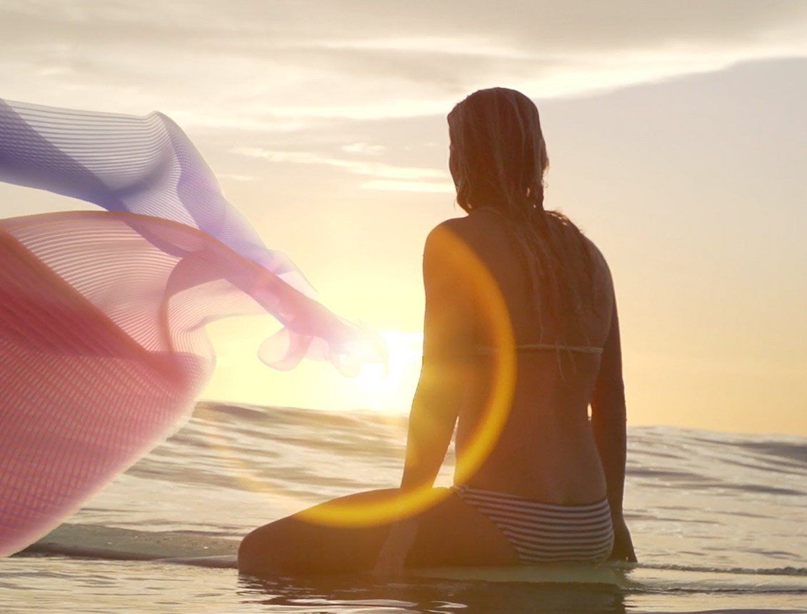 Image of women on surfboard on the ocean with a pink ribbon graphic beside her at sunset
