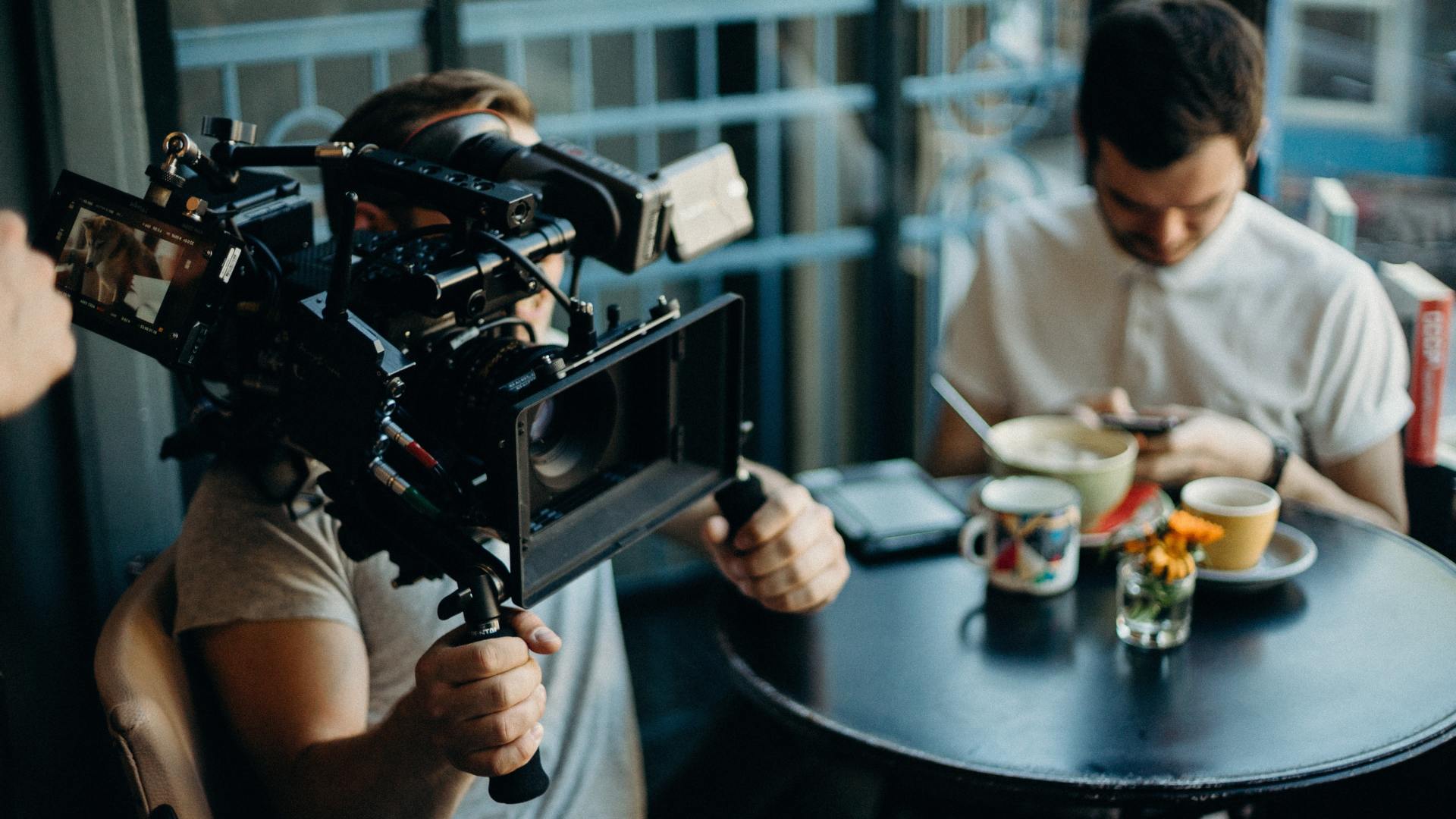 Behind the scenes image of cameraman holding a cinema camera while an actor sits in coffee shop using phone.