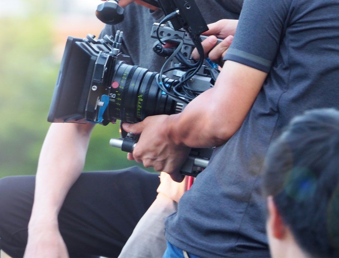 Behind the scenes photo of a video production team using a cinema camera handheld, with a large 50mm lens and matte box.