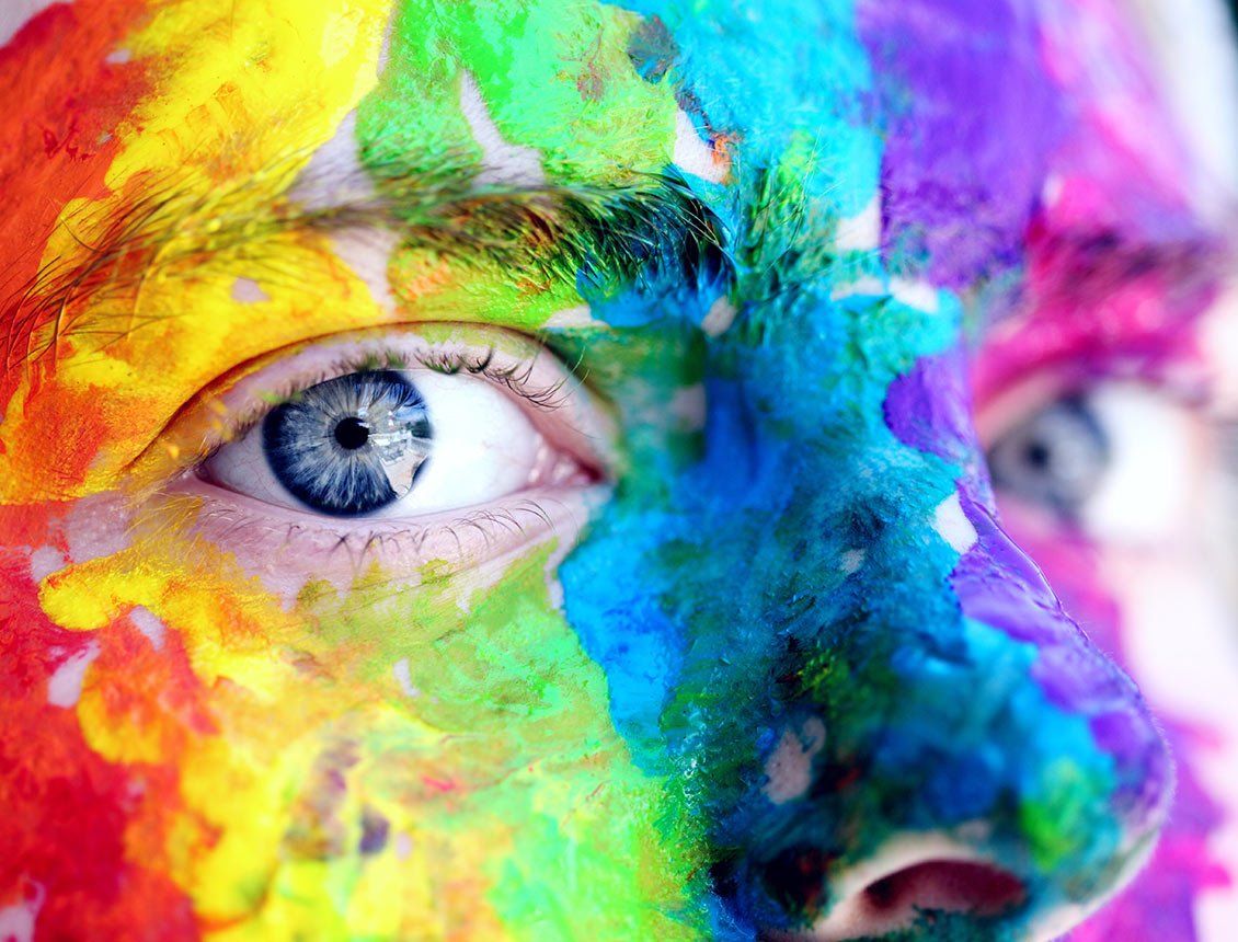 Close up image of a rainbow painted face showing a man's eye's looking into camera lens.