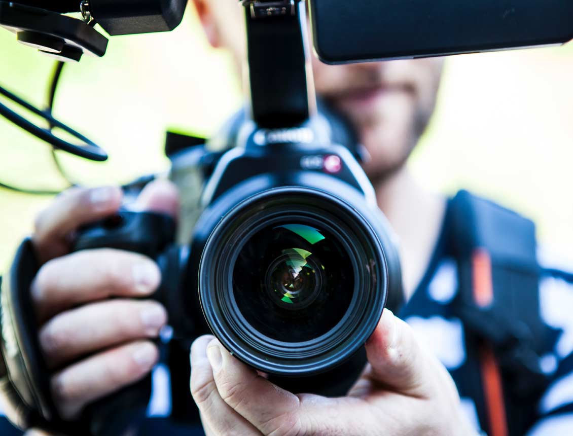 Close up image of a DSLR camera lens being held by a man. A microphone and other attachments are mounted on top of the DSLR video camera.
