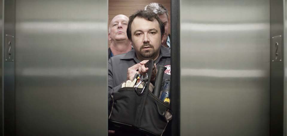 A man is standing in an elevator holding a purse.