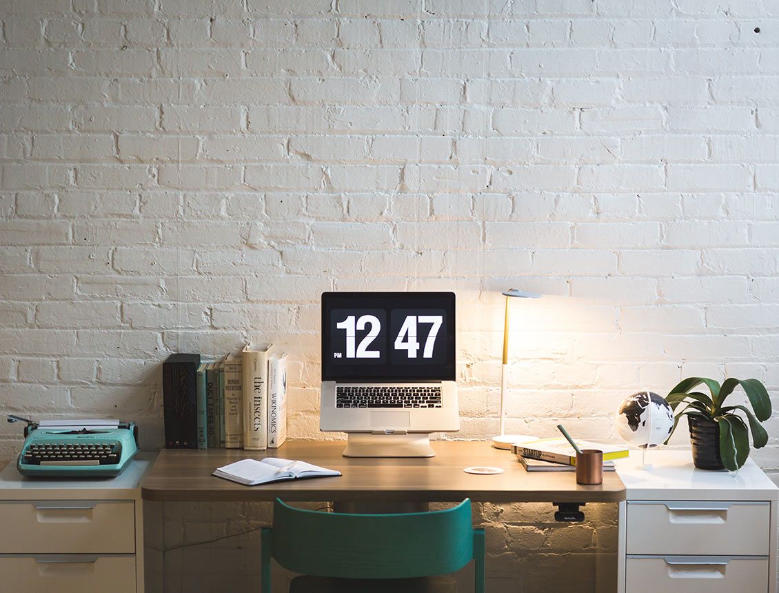 Image of a desk in front of a white brick wall with laptop that has 12:47pm on screen