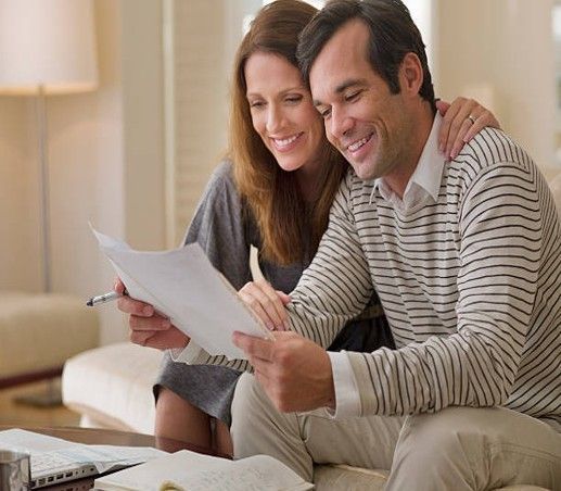 Couple smiling, reviewing paperwork together on a couch. Woman has her arm around man. Living room.