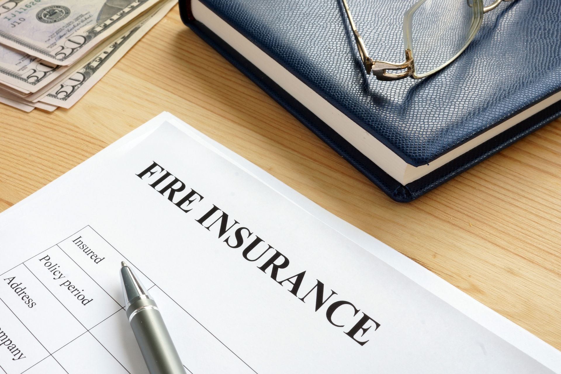 Fire insurance form, pen, notepad, glasses, and money on a wooden desk.