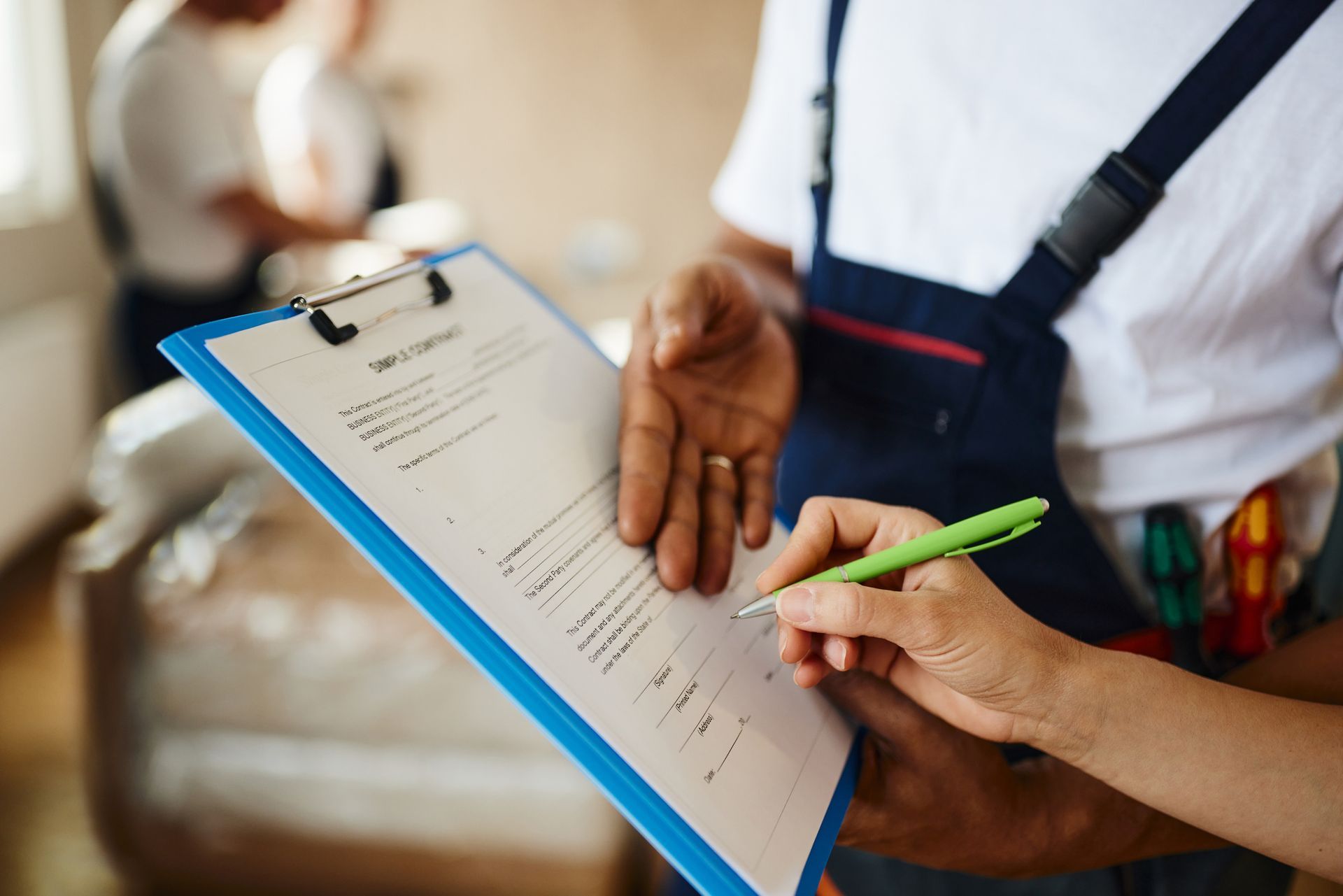 Person signing a document on a clipboard held by a worker in a white shirt and overalls, indoors. Person signing a document on a clipboard held by a worker in a white shirt and overalls, indoors.