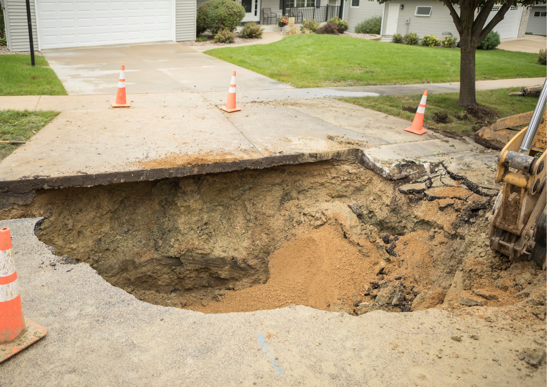 A large sinkhole in a driveway, marked with traffic cones; backhoe nearby.