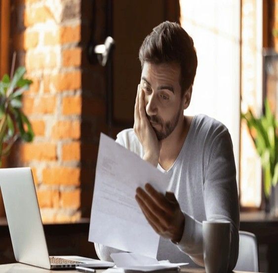 Man looking stressed while reading papers near a laptop and coffee cup; indoor setting.