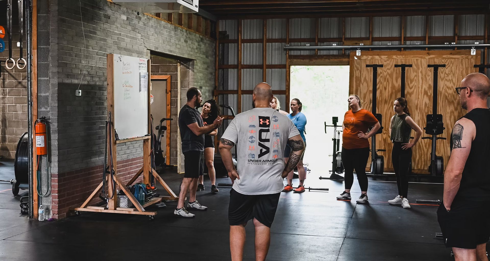 People in a gym watching a coach near a whiteboard. Several are in workout attire.