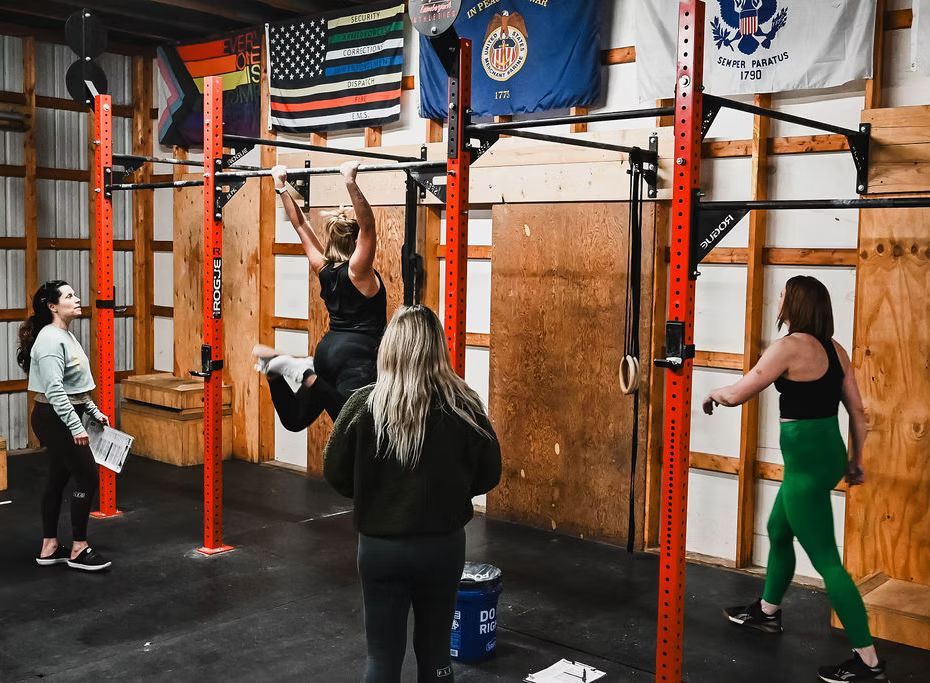 Woman doing pull-ups in a gym with other women observing. Flags and exercise equipment are visible.