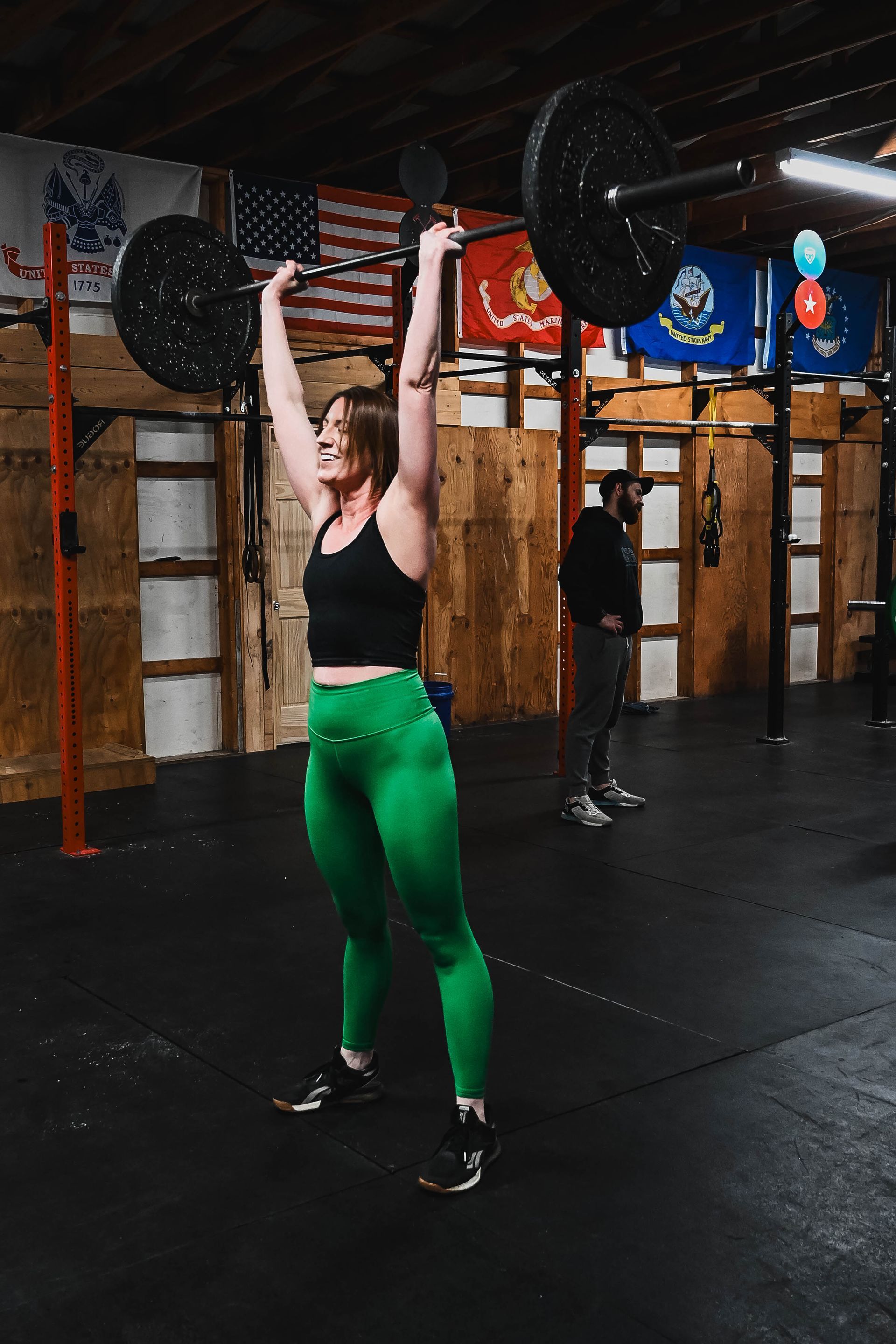 Woman lifts barbell overhead in a gym, wearing green leggings and black top; she smiles.