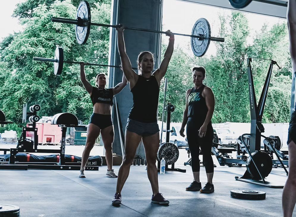 Three women doing overhead presses at a gym; one is in the center with bar raised, instructor watches.