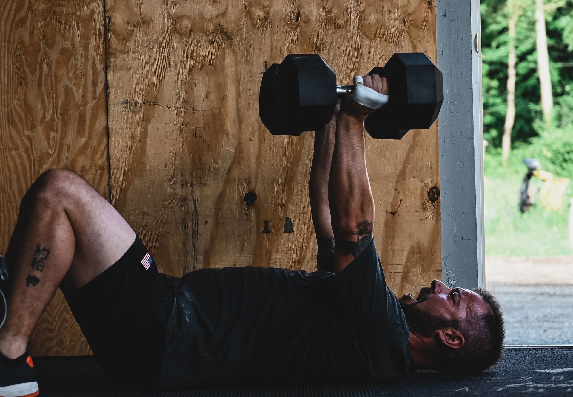 Man doing dumbbell chest press on the floor in front of a wooden wall.