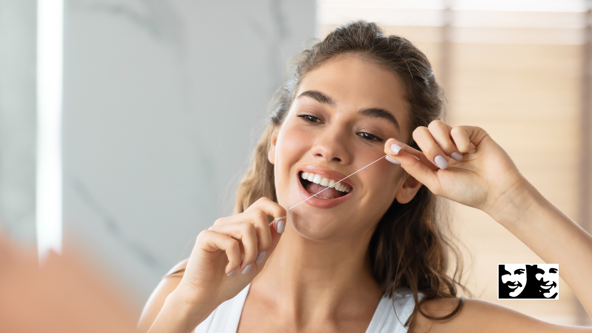 Woman flossing teeth in a bathroom, smiling. Holds floss between fingers in front of mirror.